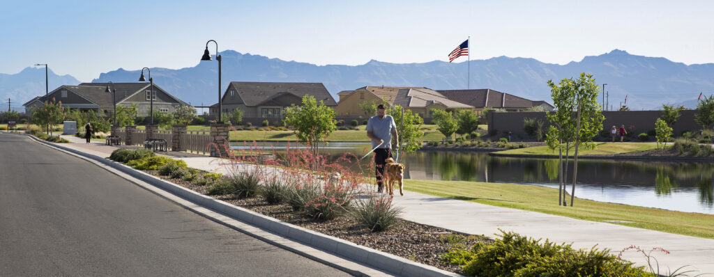 Photo of a man walking two dogs on a sidewalk near the lake. Houses and mountain views are in the background.